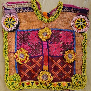 以下が含まれることがあります： ピンク、紫、オレンジの色合いの複雑な刺繍が施されたノースリーブのチュニック。黄色いビーズの縁取り、円形のビーズの花柄モチーフ、そして生地全体に小さな反射鏡のアクセントがふんだんにあしらわれた装飾的な衣服です。