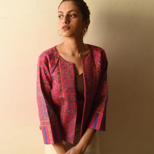 Könnte beinhalten: Eine rote, offen geschnittene Jacke mit einem Paisley-Muster in Blau, Gold und Lila. Die Jacke hat Dreiviertelärmel mit blauen und goldenen Streifen an den Manschetten. Die Jacke ist ein lebendiges Kleidungsstück.