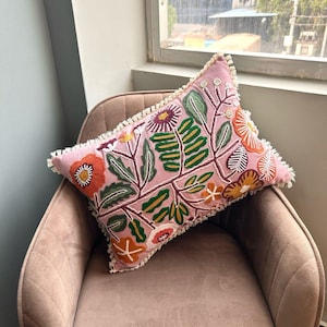 May include: A rectangular pink decorative pillow with embroidered floral designs in orange, green, and white. The pillow has a fringe trim and is placed on a pink upholstered chair. The pillow's design includes various flowers and leaves.
