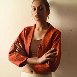 May include: A red and brown patterned kimono-style jacket with long sleeves. The jacket has a paisley design and is worn open. The model is wearing a brown top and gold hoop earrings. The jacket is a vibrant addition to any wardrobe.
