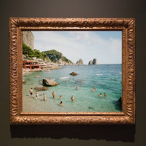 May include: A framed photograph of a beach scene with people swimming in the water. The beach is surrounded by rocky cliffs and the water is a clear turquoise blue. The photograph is framed in a gold-colored frame with ornate details.