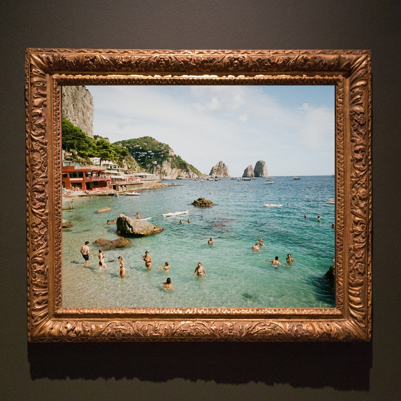 May include: A framed photograph of a beach scene with people swimming in the water. The beach is surrounded by rocky cliffs and the water is a clear turquoise blue. The photograph is framed in a gold-colored frame with ornate details.