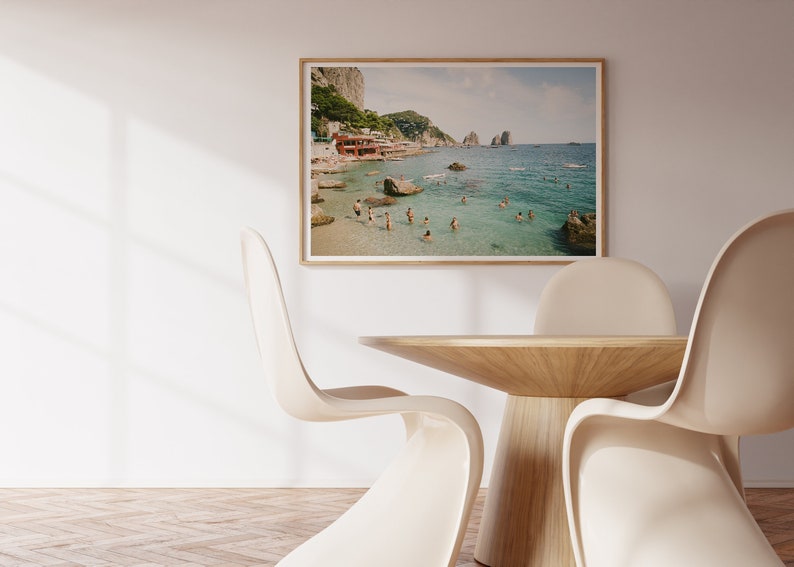 May include: A framed photograph of a beach scene with people swimming in the clear blue water. The beach is sandy with rocks and a few buildings in the background. The photo is hanging on a white wall above a wooden table with white chairs.