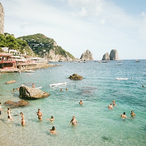May include: A beach scene with clear blue water, rocky cliffs, and people swimming and sunbathing. There are boats in the distance and a red building on the shore.