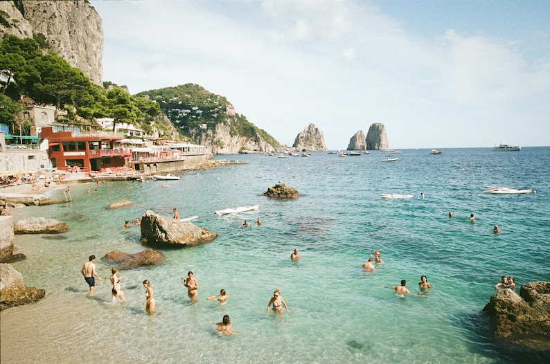 May include: A beach scene with clear blue water, rocky cliffs, and people swimming and sunbathing. There are boats in the distance and a red building on the shore.
