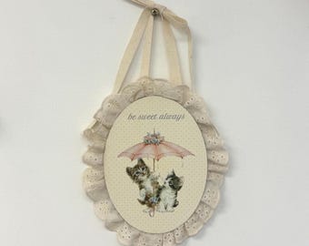 Puede incluir: Un adorno de pared ovalado con fondo color crema y un sutil patrón de puntos. En el centro, dos gatitos bajo un paraguas rosa con el texto be sweet always. La pieza está enmarcada por un borde de tela blanca con ojales fruncidos.