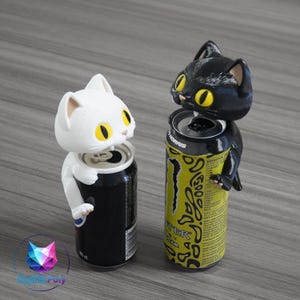May include: Two cat-shaped can toppers, one white and one black, each featuring large yellow eyes and pointed ears. The white cat sits atop a black beverage can, while the black cat sits atop a yellow and black Monster Energy drink can.