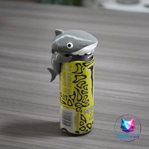 May include: A gray shark-shaped can topper sits atop a yellow and black energy drink can. The shark has white teeth and a single eye. The can has a black top and bottom with a barcode.