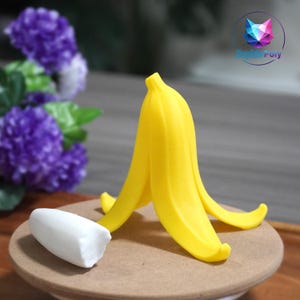 Op de afbeelding: Een heldergele bananenschil en een witte bananenschijf op een bruin oppervlak. De schil is open en toont de binnenkant. De achtergrond bevat paarse bloemen en een logo van Sophie Poly.