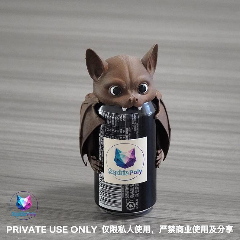 May include: A brown bat-shaped beverage can cooler featuring large eyes, pointed ears, and two small white fangs. The bat figure wraps around a black soda can with a white label displaying the text Sophie Poly and a geometric fox logo.