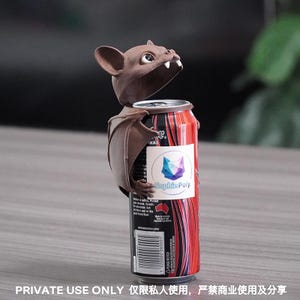 May include: A brown bat-shaped beverage can topper featuring large ears, white fangs, and outstretched wings. The topper is attached to a black and red energy drink can with a white label displaying a geometric fox logo and the text Sophie Poly.