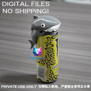 May include: A gray shark-shaped topper attached to the top of a yellow and black beverage can. The shark has a white underbelly, a single visible eye, and fins. The can features a black graphic pattern and a barcode on the lower section.
