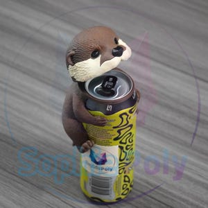 May include: A brown and white otter figurine hugging a yellow and black beverage can. The otter has a dark brown head, black nose, and white fur around its chin and neck. The can features a silver top, black pull tab, and yellow label.