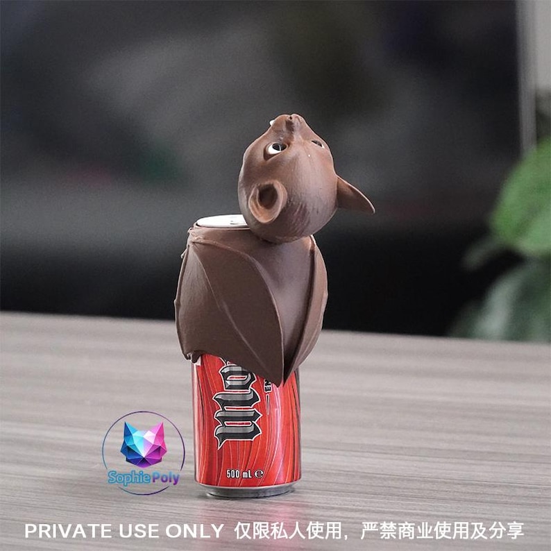 May include: A brown bat-shaped topper fitted onto a red 500 ml beverage can. The bat figure features pointed ears, visible eyes, and folded wings that wrap around the top portion of the can. The can displays the text "Monster" in white font.