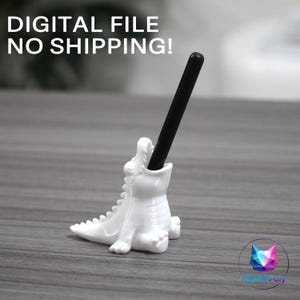 May include: A white, stylized alligator-shaped pen holder with a black pen inserted into its open mouth. The text DIGITAL FILE NO SHIPPING! is visible at the top. The object features a textured back with ridges and a smooth, glossy finish.