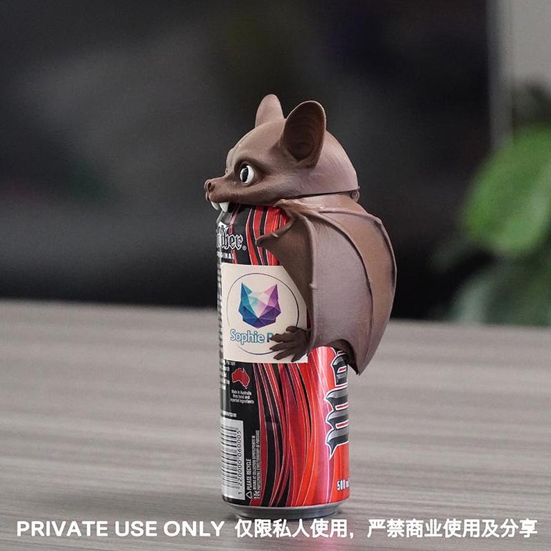 May include: A brown bat-shaped beverage can topper featuring large ears, visible white fangs, and folded wings. The topper is attached to a red and black 500 ml beverage can with a label displaying the text Sophie P and a geometric fox logo.