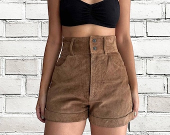 Op de afbeelding: Bruine shorts met hoge taille, gerolde zoom en sluiting met twee knopen. De shorts hebben voorzakken en een brede tailleband. Het model draagt ook een zwarte top met spaghettibandjes.