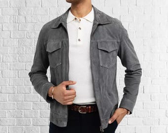 Può includere: Giacca in camoscio grigio con due tasche frontali e chiusura con zip. La giacca è indossata sopra una polo bianca e una cintura in pelle marrone. Lo sfondo è un muro di mattoni bianchi.