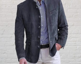 Puede incluir: Un blazer de ante gris carbón oscuro con cuello mao, cierre frontal de botones y dos bolsillos con solapa en la cintura. El blazer se lleva sobre una camisa de rayas verticales azules y blancas y unos pantalones de color claro.