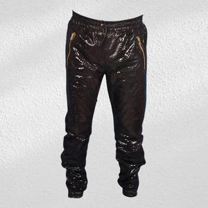 Puede incluir: Pantalones negros de piel sintética con estampado de piel de serpiente. Los pantalones tienen cremalleras doradas en los bolsillos y cintura con cordón. Los pantalones están fruncidos en los tobillos.