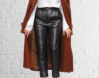 Op de afbeelding: Een lange, roestkleurige jas met een knoopsluiting, gedragen over een witte blouse en een zwarte leren broek. De broek heeft een hoge taille en is getailleerd. De outfit staat tegen een witte bakstenen muur.