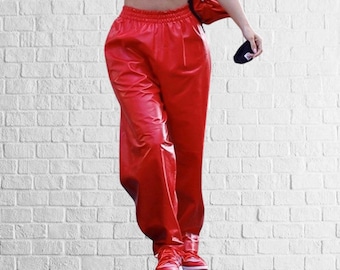 May include: Vibrant red, shiny trousers with an elasticated waistband and drawstring. The trousers are worn with matching red trainers with white soles and laces. The background is a white brick wall.