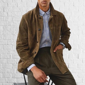 以下が含まれることがあります： ボタン留めの襟付きブラウンのスエードジャケット。明るいブルーのボタンダウンシャツと濃い緑色のパンツの上に着用。左手首には腕時計が見える。