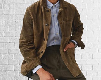 May include: A brown suede jacket with a button-up front and a collared neckline. The jacket is worn over a light blue button-down shirt and dark green pants. The person is wearing a watch on their left wrist.