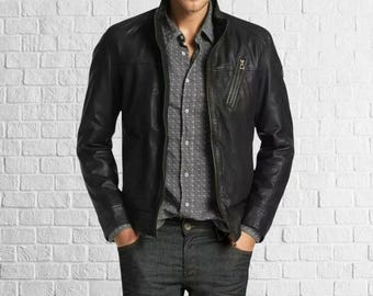 May include: A black leather jacket with a zip closure and a stand-up collar. The jacket is worn over a gray button-down shirt with a subtle pattern. The person is wearing dark wash jeans. The background is a white brick wall.
