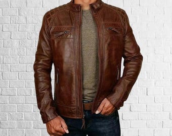 May include: A brown leather jacket with a stand-up collar and diamond-stitched shoulder details. The jacket has multiple zippered pockets and a front zipper closure. The model is wearing a gray shirt and blue jeans.