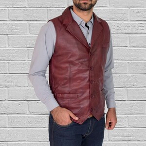 Puede incluir: Un chaleco de cuero burdeos con solapa de muesca y cierre de tres botones. El chaleco tiene dos bolsillos delanteros y un corte entallado. El modelo lleva una camisa gris y vaqueros azules. El fondo es una pared de ladrillos blancos.