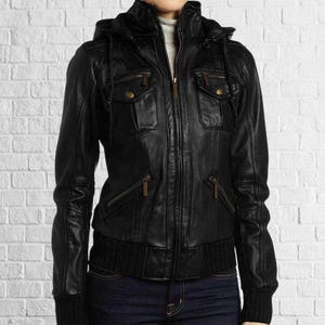 Puede incluir: Chaqueta de cuero negra con capucha, m&uacute;ltiples bolsillos y cierre de cremallera. La chaqueta tiene un dobladillo y pu&ntilde;os de punto acanalado. La chaqueta se usa sobre un cuello alto gris y jeans azules.