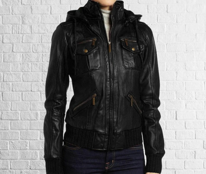 Puede incluir: Chaqueta de cuero negra con capucha, m&uacute;ltiples bolsillos y cierre de cremallera. La chaqueta tiene un dobladillo y pu&ntilde;os de punto acanalado. La chaqueta se usa sobre un cuello alto gris y jeans azules.