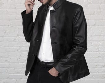 Puede incluir: Una chaqueta de cuero negra con cuello mao y cierre de botones en la parte delantera. La chaqueta se lleva sobre una camisa blanca de botones y se combina con pantalones negros y un cinturón de cuero negro, logrando un aspecto estructurado.