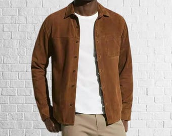 May include: A brown suede shirt with a button-down front and a single chest pocket. The shirt is worn over a white t-shirt and paired with khaki pants. The background is a white brick wall.