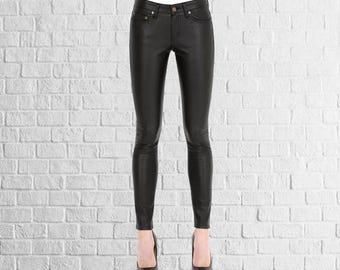 Op de afbeelding: Zwarte broek met leerlook en een klassiek ontwerp met vijf zakken. De broek is nauwsluitend en heeft een knoop- en ritssluiting. Het model draagt zwarte hoge hakken. De achtergrond is een witte bakstenen muur.