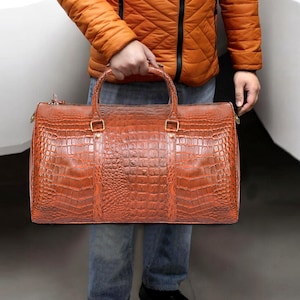 May include: A brown leather duffel bag with a crocodile skin texture. The bag has two handles and a zipper closure. The bag is being held by a person wearing a burnt orange jacket and blue jeans.