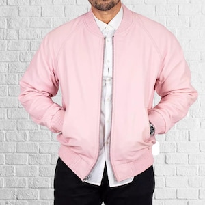 Op de afbeelding: Een lichtroze bomberjack met een geribbelde kraag, manchetten en zoom. De jas heeft een ritssluiting en twee voorzakken. Daaronder wordt een wit overhemd met knopen gedragen. De achtergrond is een witte bakstenen muur.