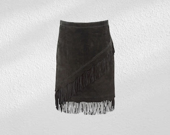 May include: A dark gray suede skirt with a fringed diagonal panel. The skirt has a wrap-style design and a straight hemline. The fringe detail adds a bohemian flair to the garment. The skirt is made of a soft, textured material.