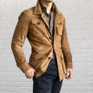 Könnte beinhalten: Eine hellbraune Wildlederjacke mit Kragen, durchgehender Knopfleiste und zwei Brusttaschen mit geknöpften Klappen. Die Jacke hat lange Ärmel mit geknöpften Manschetten und wird über einem dunkelgrauen Hemd und einer dunklen Hose getragen.