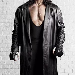 May include: A long black leather coat, open to reveal a black vest and gloves. The image is set against a white brick wall. The coat's length is notable.