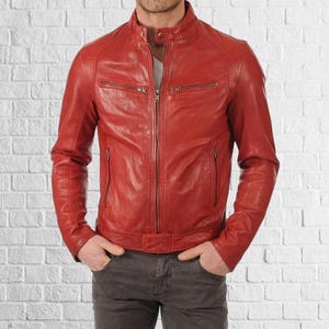 May include: A red leather jacket with a zip-up front, two chest pockets, and two side pockets. The jacket has a stand-up collar and is worn over a white shirt. The model is wearing gray pants.
