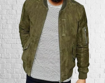 May include: Olive green suede bomber jacket with a zip closure, side pockets, and a ribbed collar and cuffs. The jacket is worn over a black and white striped t-shirt. The background is a white brick wall.