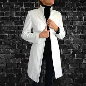 Op de afbeelding: Een witte, lange jas met een hoge kraag en een getailleerde pasvorm. De jas is aan de voorkant open en onthult een zwarte top en broek. De achtergrond is een donkere bakstenen muur.
