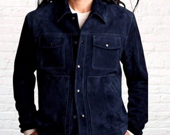 Può includere: Una giacca in camoscio blu navy scuro con colletto classico, chiusura frontale con bottoni metallici, due tasche sul petto con patta abbottonata e due tasche laterali inferiori. La giacca è indossata sopra una camicia bianca semplice.