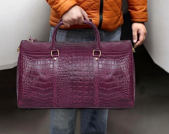May include: A deep purple duffel bag with a crocodile skin texture. The bag has two handles and gold-colored hardware. The person holding the bag is wearing blue jeans and a bright orange jacket.