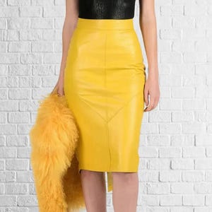 May include: A bright yellow leather pencil skirt with a high waist and a knee-length hem. The skirt has a unique geometric design on the front. A fluffy yellow jacket is held in the left hand. The top is black.