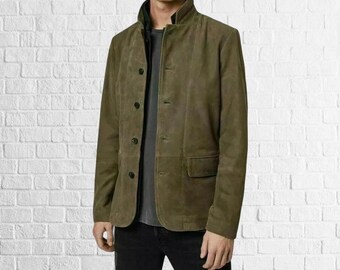 May include: Olive green suede jacket with a button-up front, a small pocket, and a stand-up collar. The jacket is worn over a dark gray shirt and black pants. The background is a white brick wall.
