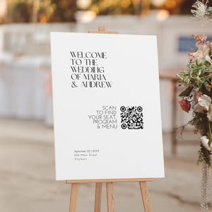 Op de afbeelding: Een wit trouwbord op een houten ezel. Het bord toont zwarte tekst: "WELCOME TO THE WEDDING OF MARIA & ANDREW." Een QR-code en de woorden "SCAN TO FIND YOUR SEAT, PROGRAM & MENU" staan eronder. De datum en het adres zijn ook afgedrukt.