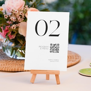 Op de afbeelding: Een witte tafelnummerkaart met het nummer "02" in grote zwarte letters. De kaart bevat ook de tekst "PROGRAMMA & MENU", een QR-code en namen met een datum. De kaart wordt weergegeven op een kleine houten ezel.